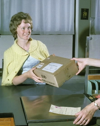 Eva Sandström (fingerat namn) hämtar ett paket i postluckan mot överlämnande av det kvitterade adresskortet, som postkassören tar emot. Den arrangerade situationen är fotograferad i paketkassan på postkontoret Farsta 1.